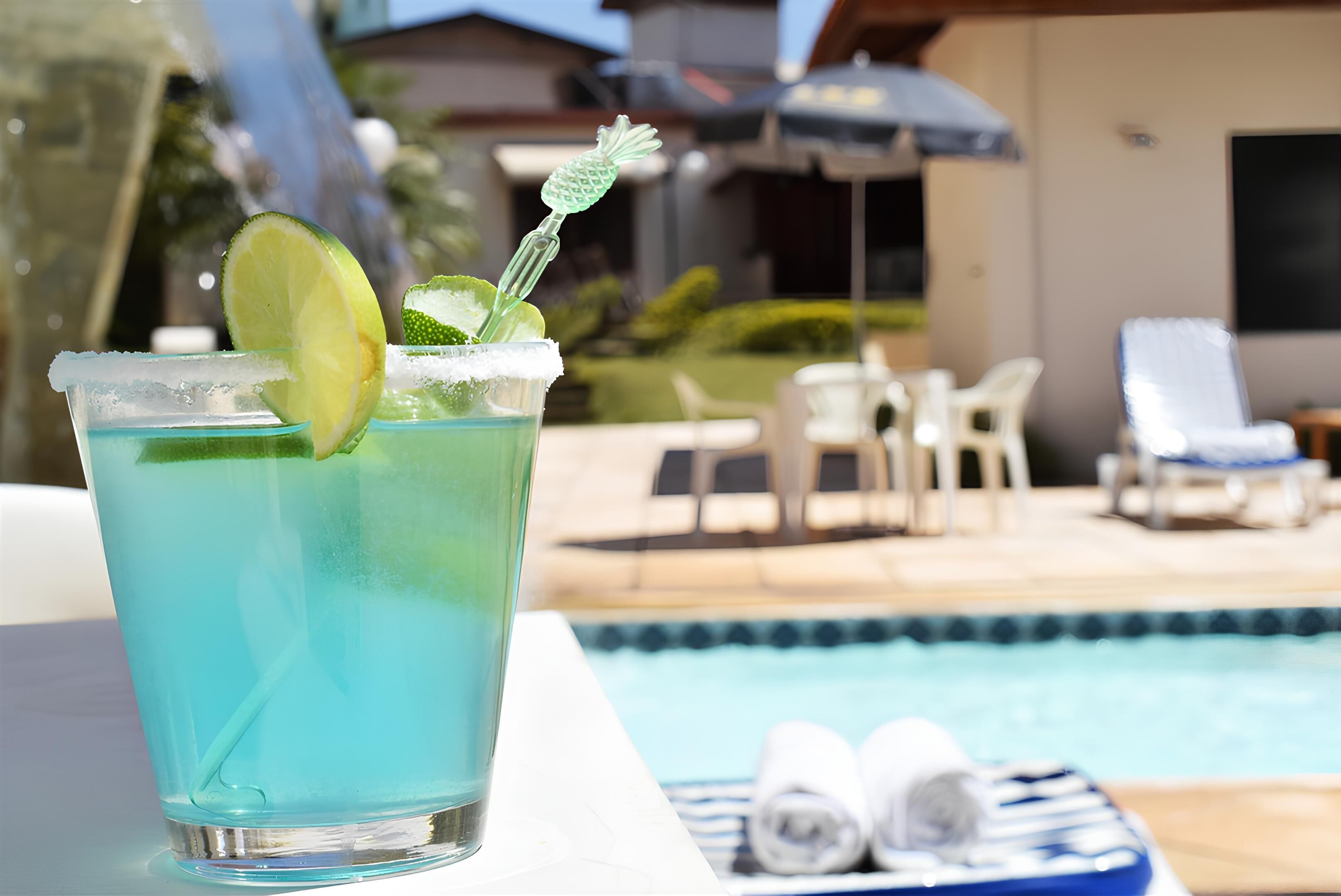 Un verre transparent contenant une boisson bleue, une tranche de citron vert et une feuille de menthe en guise de décoration, posé sur une table près d’une piscine, avec des chaises longues et des parasols.