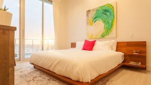 Un dormitorio moderno con una cama grande, cabecera de madera, vistas de la ciudad a través de amplios ventanales y un cuadro en la pared.