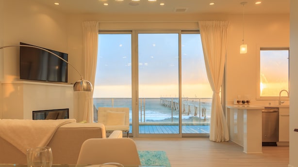 Habitación de hotel con un amplio ventanal corredera que da a un balcón con vistas al océano y un muelle. Cuenta con un comedor con una mesa para dos, un televisor montado en la pared y una zona de cocina con fregadero y encimera.