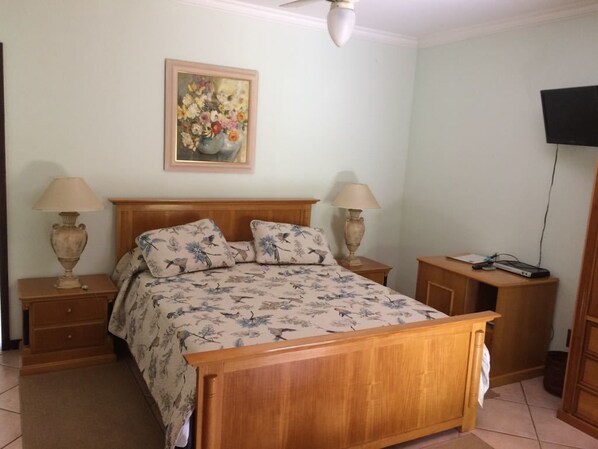 A bedroom with a wooden bed, two bedside tables with lamps, a painting on the wall, and a TV on the right.