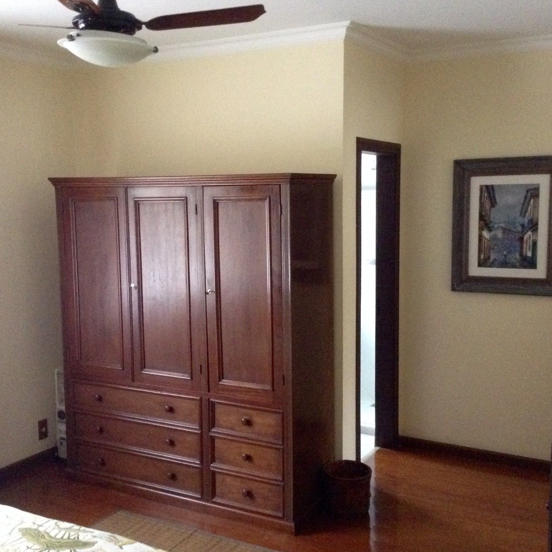 Une chambre avec une grande armoire en bois, un ventilateur de plafond et un tableau encadré accroché au mur.