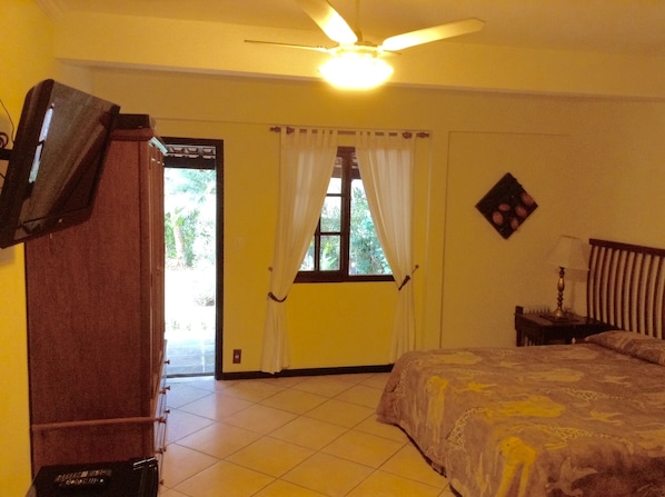 Una habitación con una cama, un ventilador de techo, un televisor montado en la pared y una ventana con cortinas.