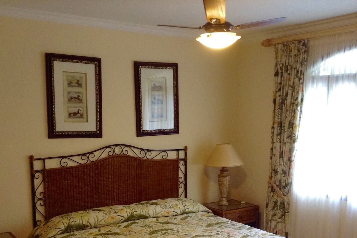 A neatly made bed with a floral bedspread, a wooden headboard, two framed pictures on the wall, a bedside lamp, and a window with curtains.