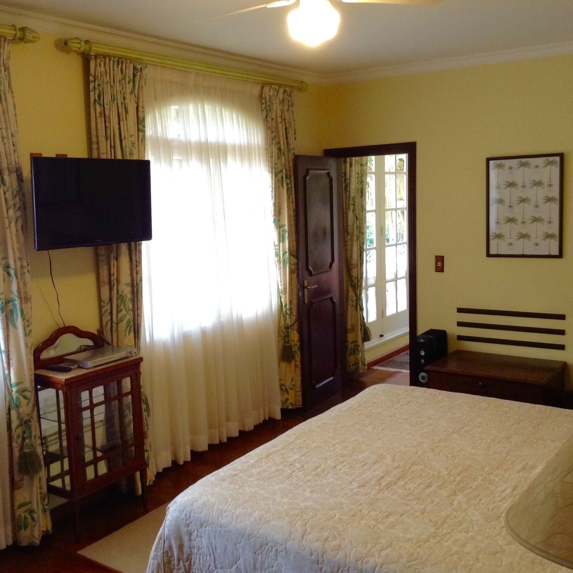 A bedroom with a bed, a television on the wall, a desk with a laptop, and a door leading to another room.