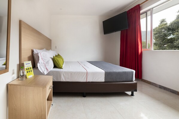 A bedroom with a bed, a wooden nightstand, a bottle, a box, a TV, and a window with red curtains. - Ayenda 1238 Nuevo Colombia (Medellín)
