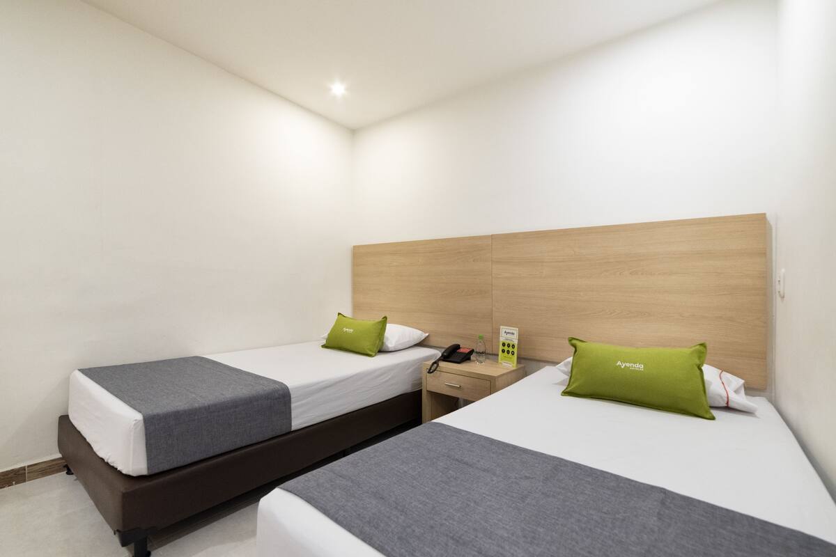 A hotel room with two beds, each with a grey bedspread and a green pillow. There is a wooden headboard, a bedside table with a phone and a small box, and a wall-mounted light.