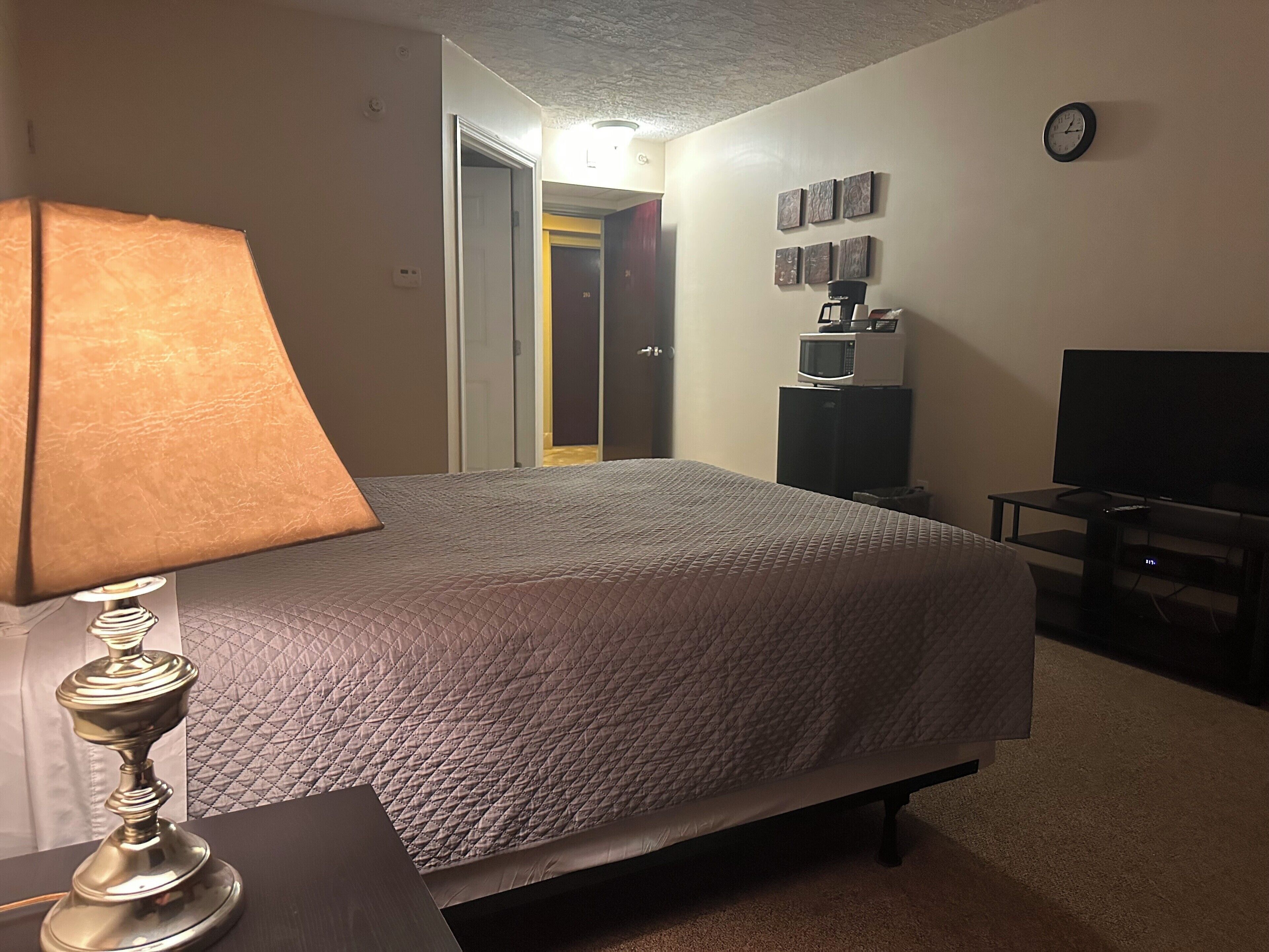 A hotel room with a bed, a lamp, a television, a coffee machine, and a clock on the wall.