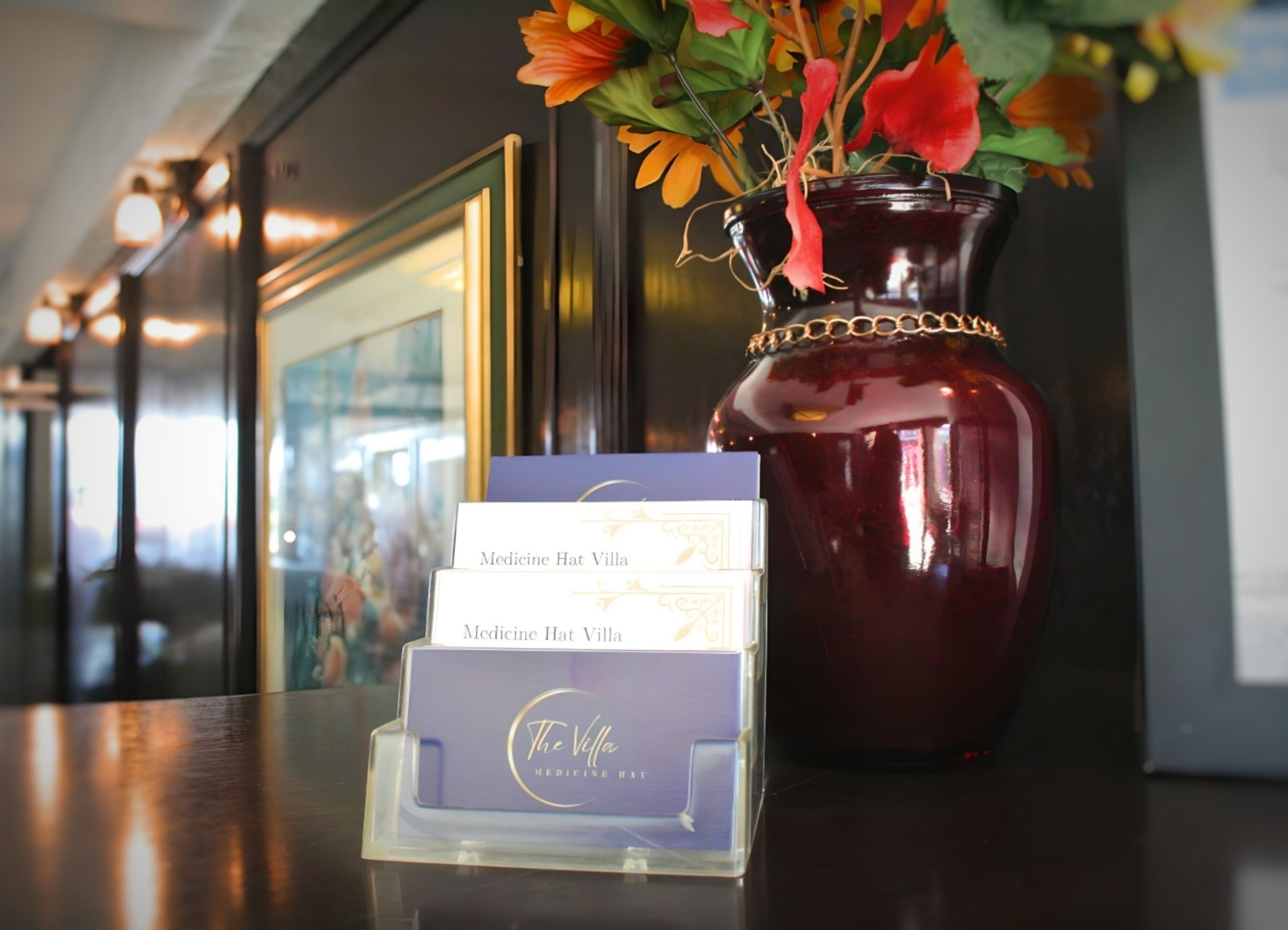 A dark red vase with a gold chain, filled with colorful flowers, and a display stand with 'Medicine Hat Villa' branded cards.