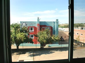 Blick durch ein Fenster auf ein Gebäude mit Wandgemälde, Bäume und blauem Himmel.