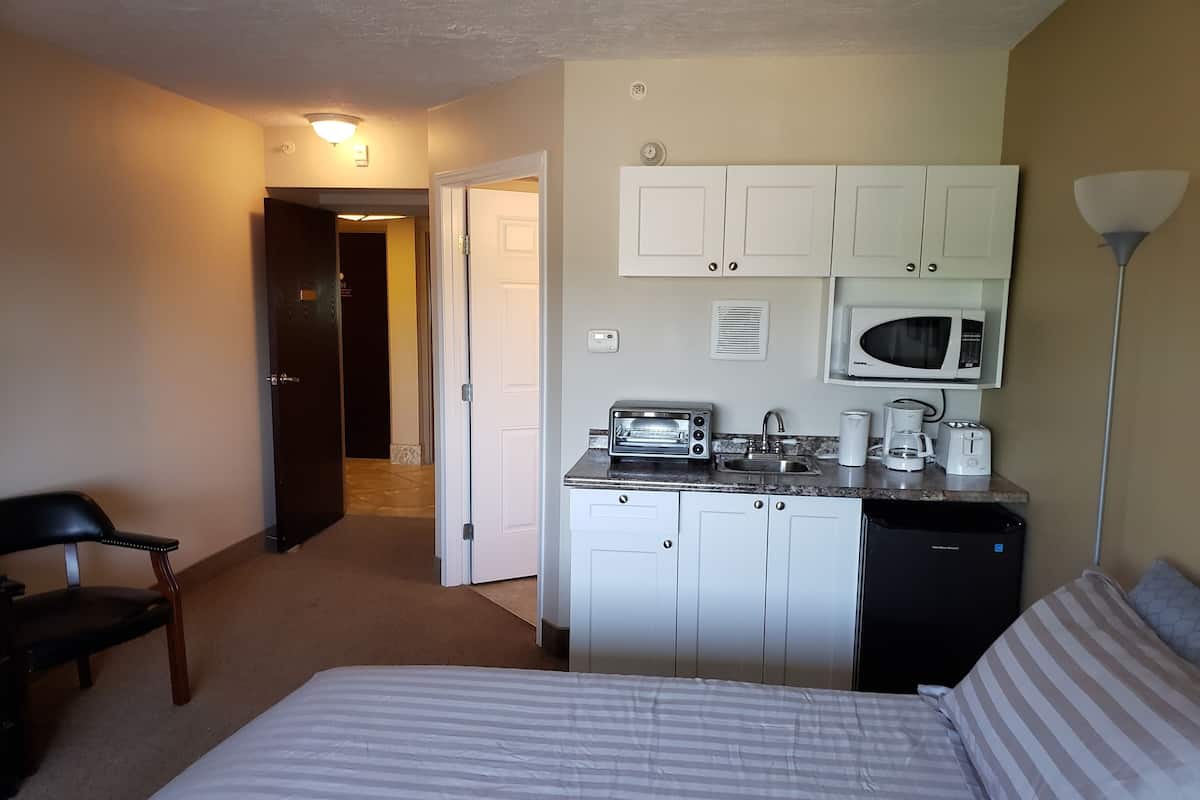 Een compacte hotelkamer met kitchenette, een bed met gestreept beddengoed, een stoel en een deur naar een andere kamer.