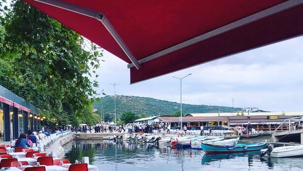 A waterfront dining area with tables and chairs under a red canopy, boats docked in the background, and a view of the harbor. - Adasu Otel (Marmara)