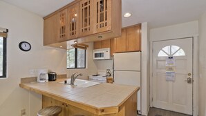 Una cocina con armarios de madera, microondas, refrigerador, pileta y dos taburetes.