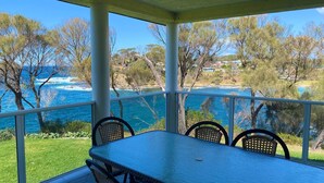 Un espacio de estar al aire libre cubierto con mesa y sillas, con vistas a un paisaje costero con árboles y el mar.