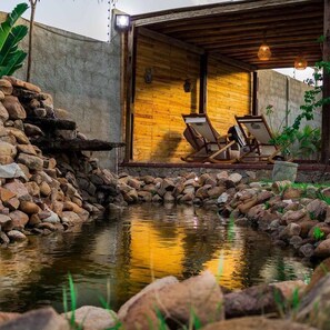 Una cabaña rústica con pared de piedra, una terraza de madera y un pequeño estanque.
