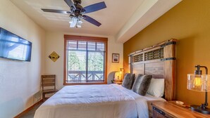Ein Schlafzimmer mit einem Bett, einem Nachttisch aus Holz, einer Schreibtischlampe, einem Deckenventilator, einem Fenster mit Jalousien, einem Stuhl und einem an der Wand montierten Fernseher.