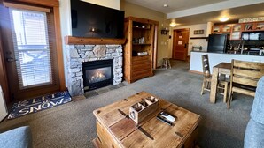 Ein gemütliches Wohnzimmer mit Kamin aus Stein, Holzmöbeln und einer Küchenzeile mit schwarzem Kühlschrank.