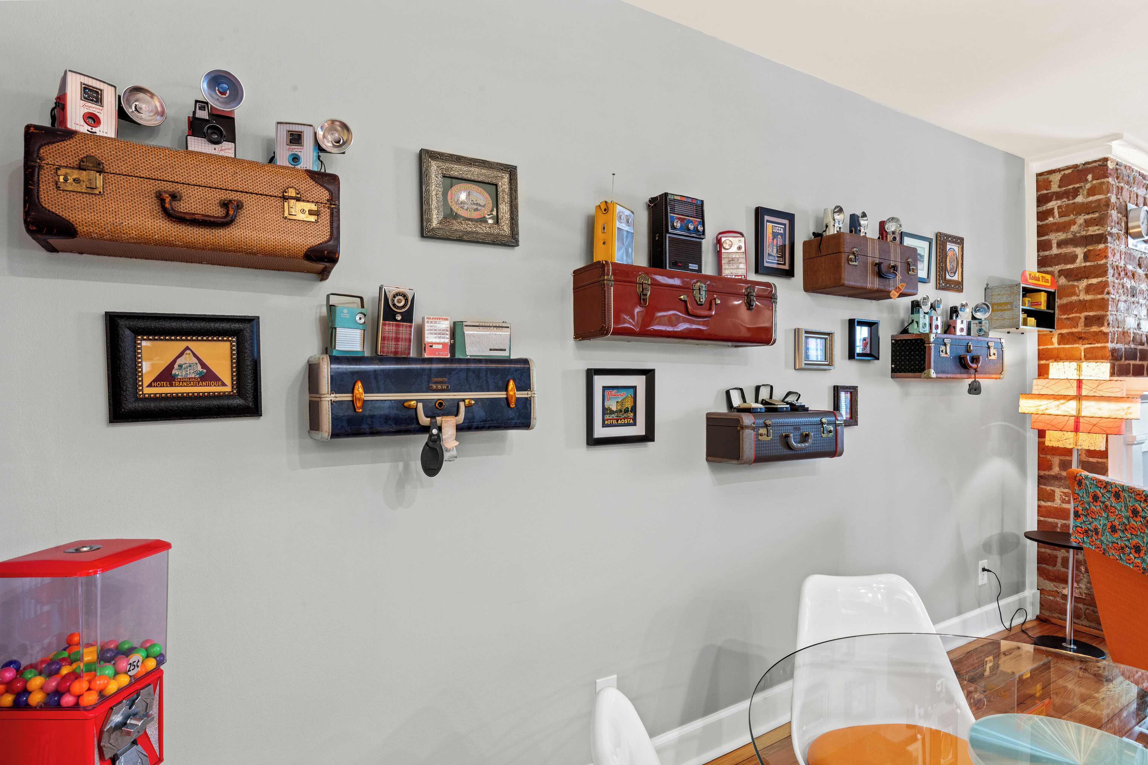 A wall with various vintage suitcases and cameras, a red vending machine with colorful candies, and a glass table with a clear glass top.