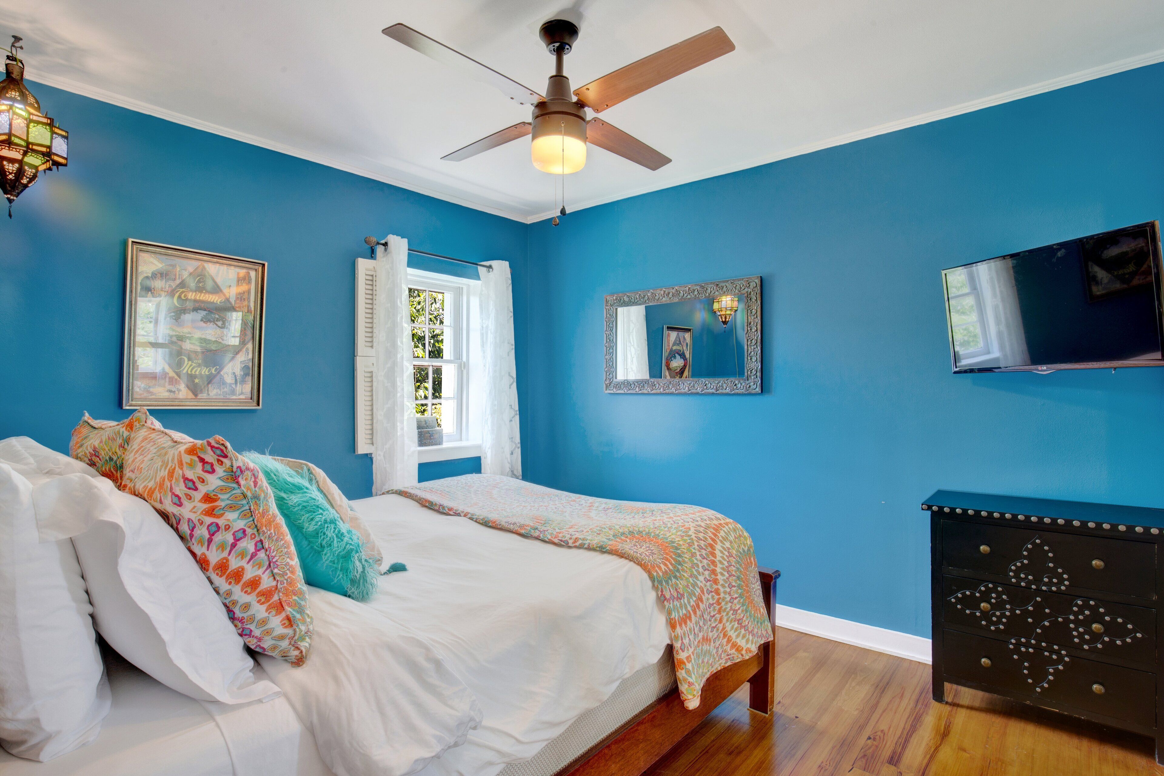 A bedroom with a bed, a ceiling fan, a dresser, a TV, and a window with curtains.