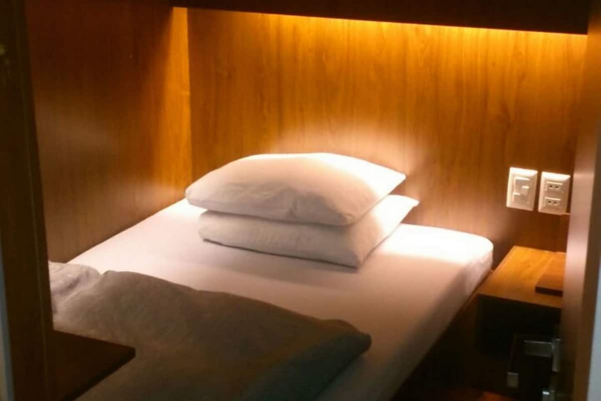 A compact, wooden bunk bed with a single pillow and a blanket, a bedside table with a lamp, and a power outlet.