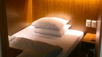 A compact, wooden bunk bed with a single pillow and a blanket, a bedside table with a lamp, and a power outlet.