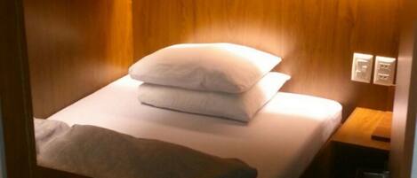 Una cama nido de madera compacta con una almohada y una colcha, una mesita de noche con una lámpara y un enchufe.