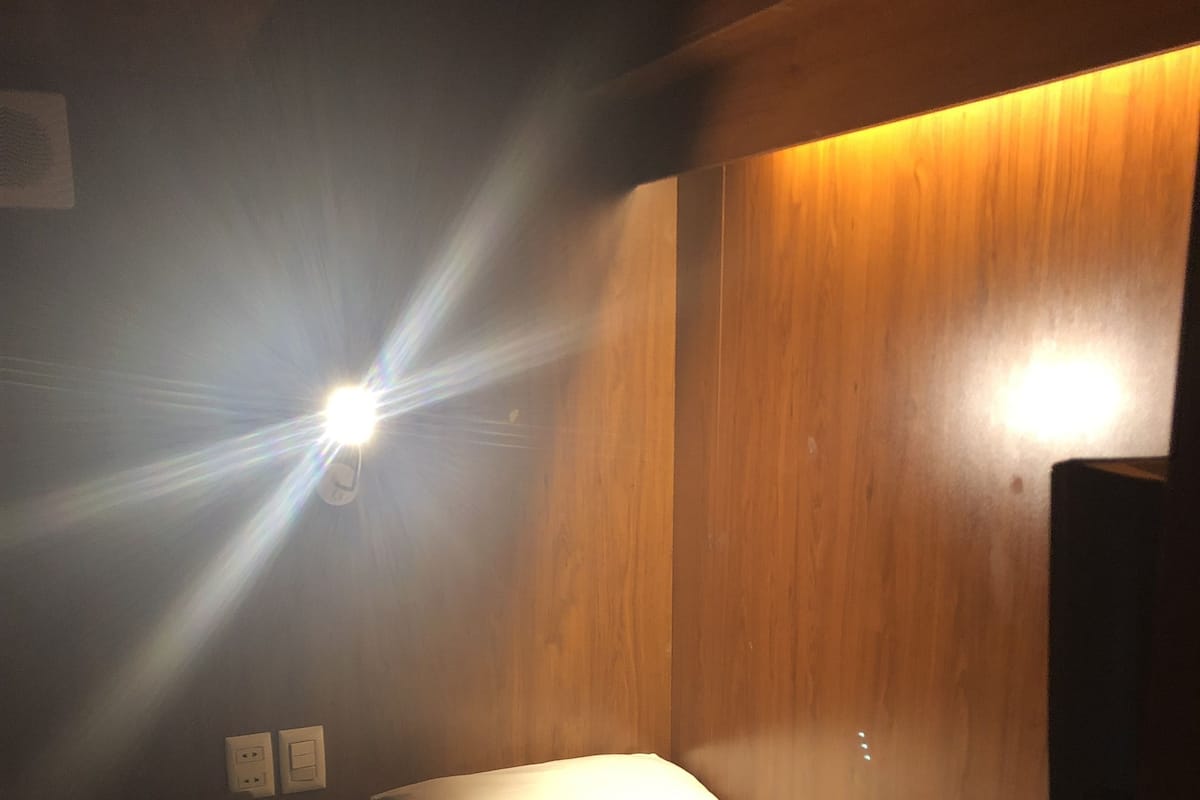 A small, dimly lit room with a single bed, a wooden headboard, and a wall-mounted light.