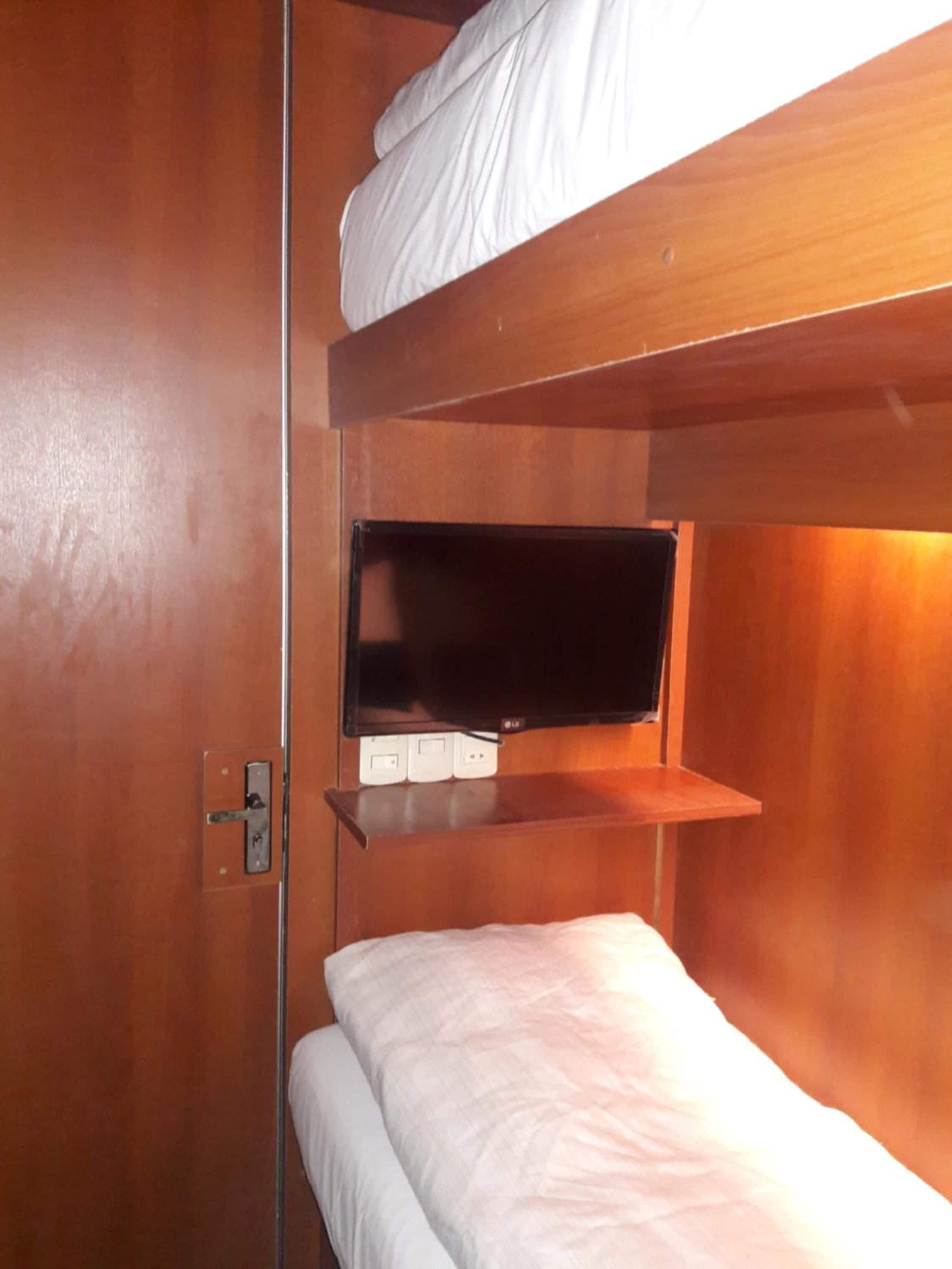 Una cama litera con una cama baja y un estante arriba, en una habitación con paredes de madera y un enchufe a la vista.