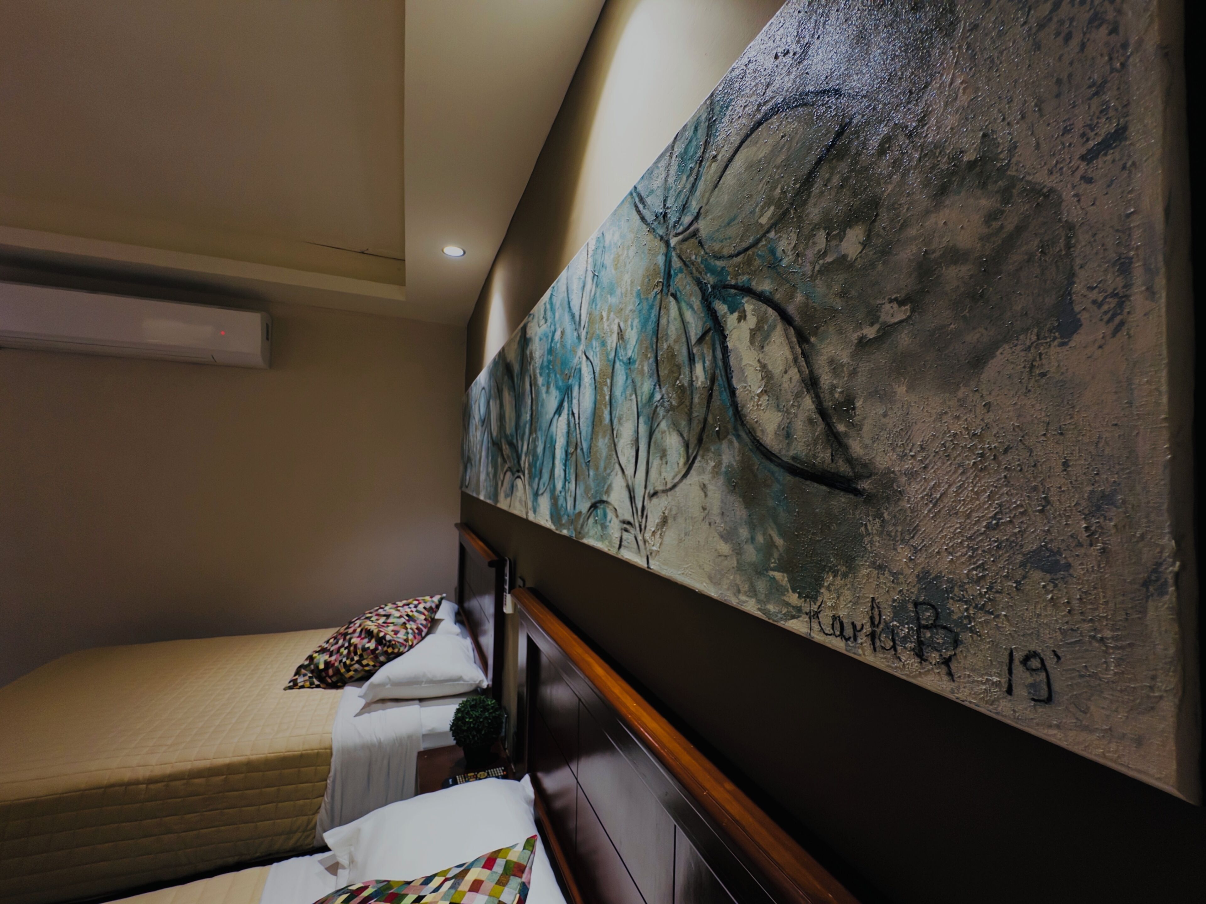 Habitación de hotel con cama, un cuadro abstracto grande en la pared y una lámpara de techo.