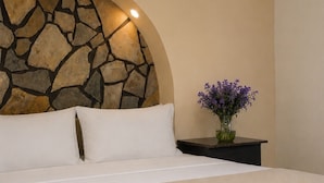 Un letto con lenzuola bianche, una testiera in pietra, un vaso con fiori viola e un tavolino accanto al letto.