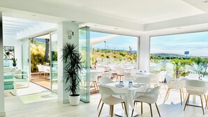 Un espace de restauration moderne avec des chaises et des tables blanches, de grandes fenêtres donnant sur un jardin aménagé et des collines.