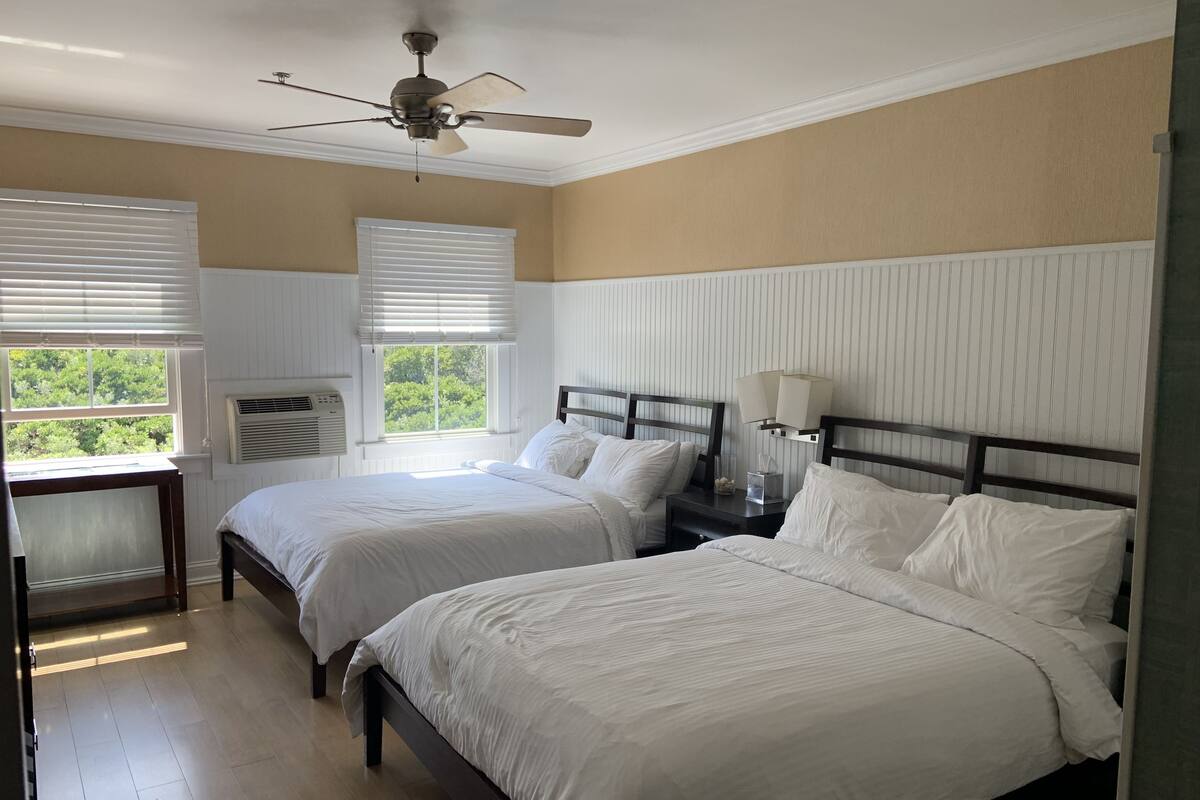 A hotel room with two beds, a ceiling fan, a window with blinds, an air conditioning unit, and a bedside table with a lamp.