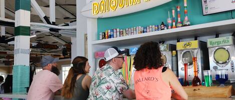 Un bar anomenat "Daiquiri Bar" amb un rètol de tabla de surf, diverses begudes a les estanteries i un tauler de preus.