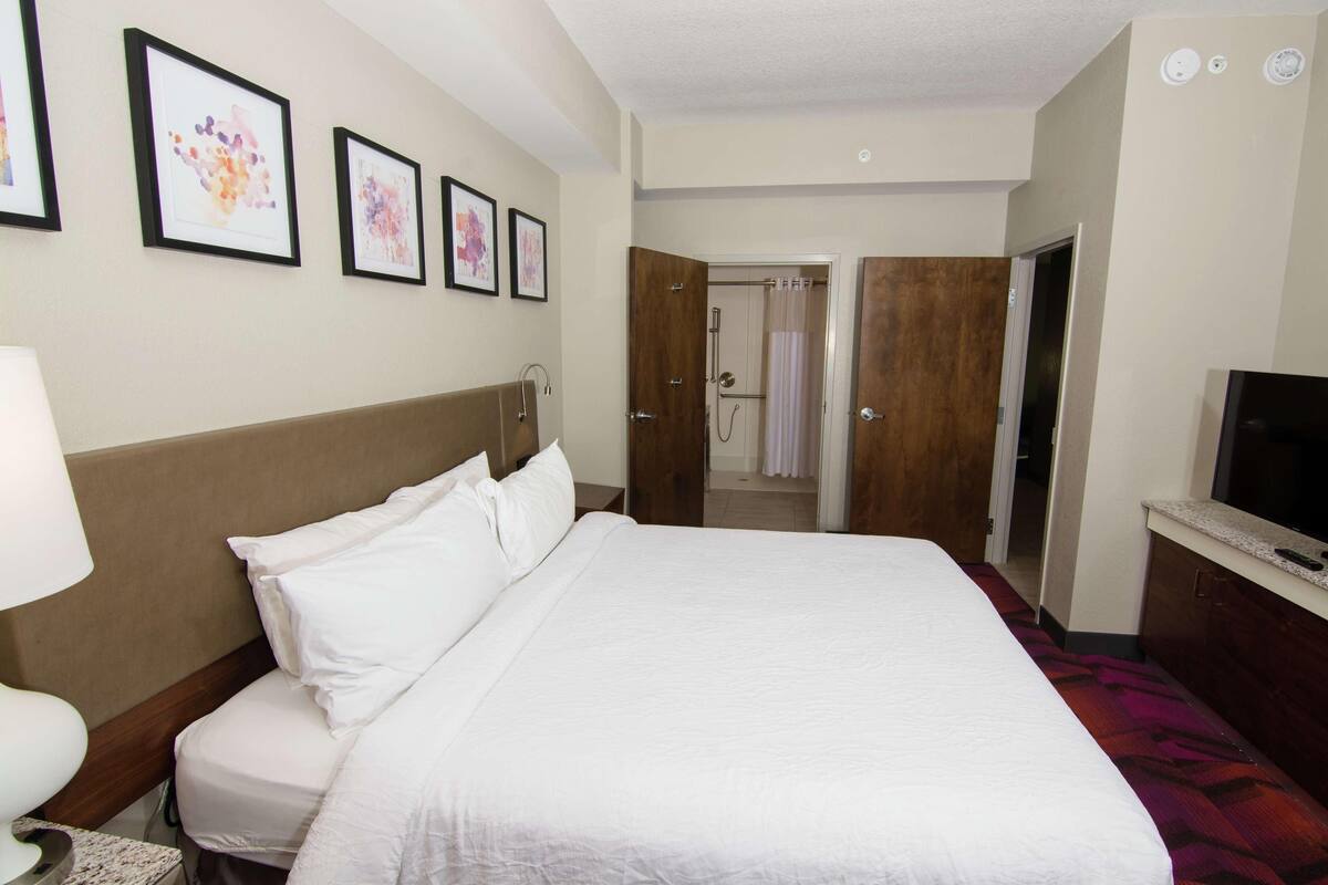 A hotel room with a bed, a TV, a bathroom with a shower curtain, and framed artwork on the wall.