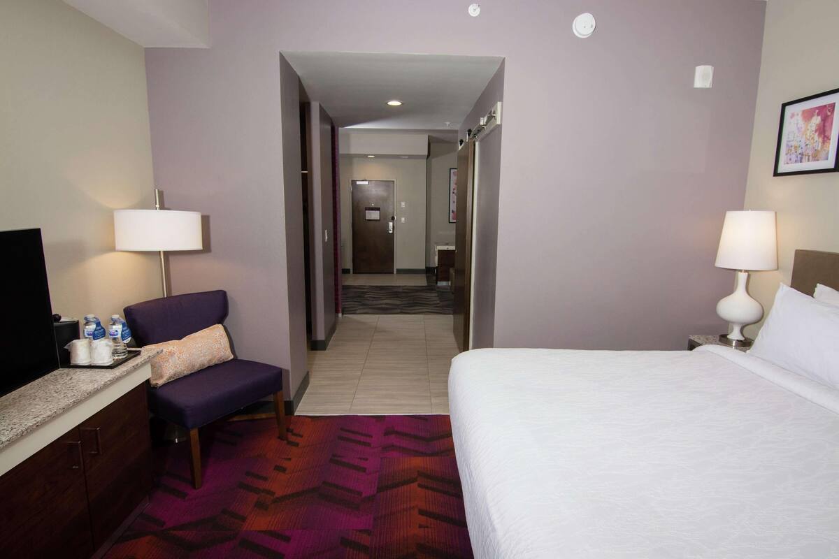 A hotel room with a bed, a chair, a desk with a TV, and a bathroom visible through an open door.