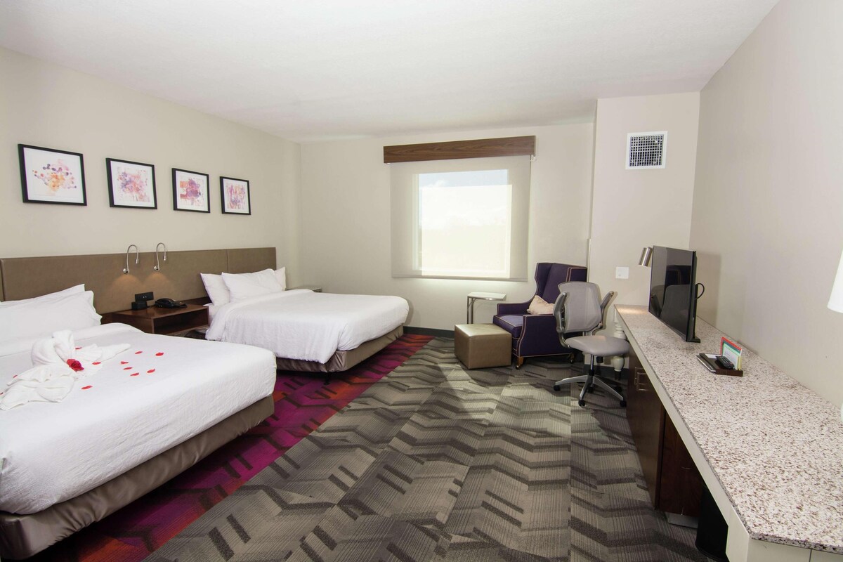 A hotel room with two beds, a desk, a chair, a TV, and a window.