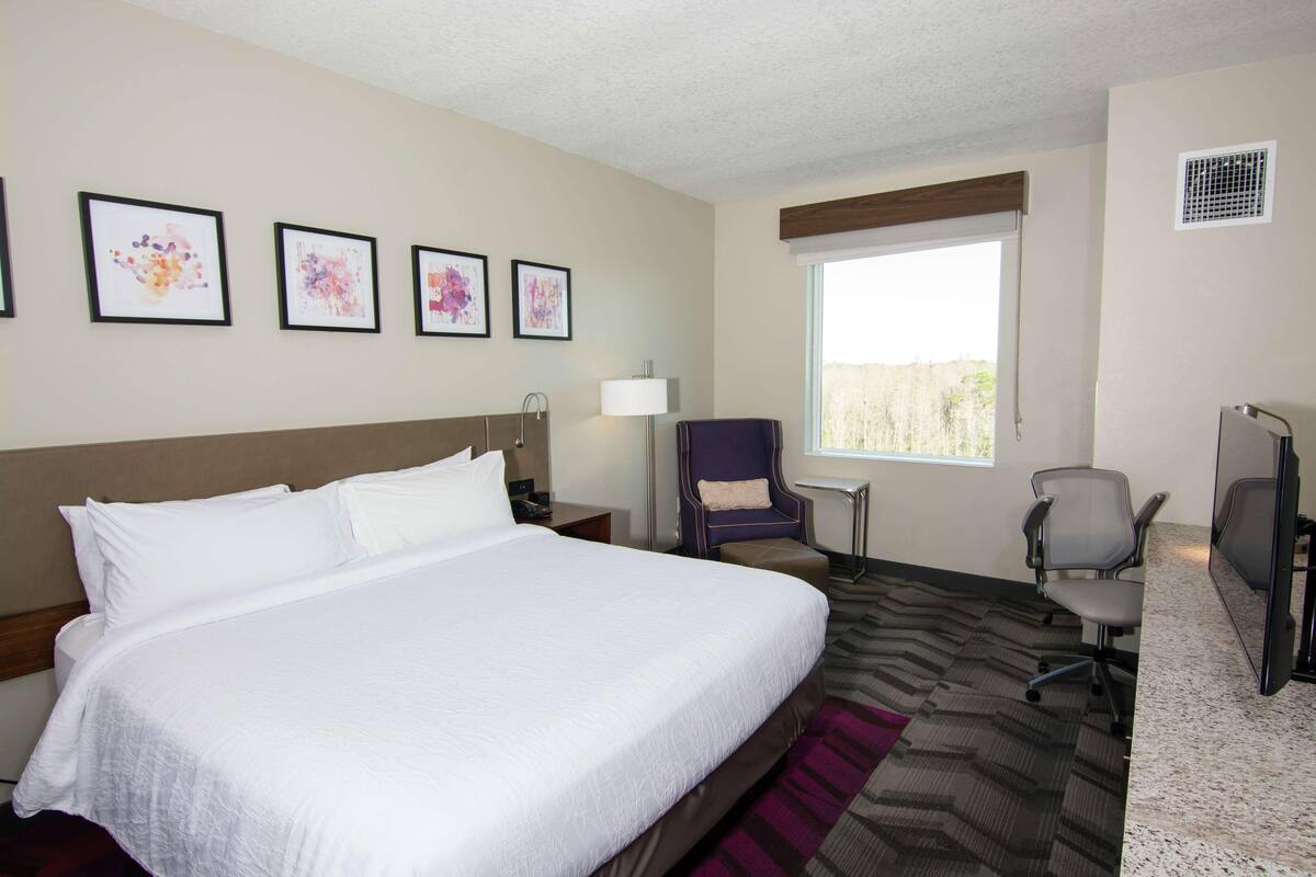 A hotel room with a large bed, a desk with a chair, a window, and artwork on the wall.