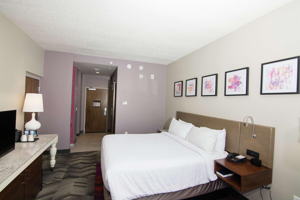 A hotel room with a bed, bedside table, lamp, TV, and framed artwork on the wall.