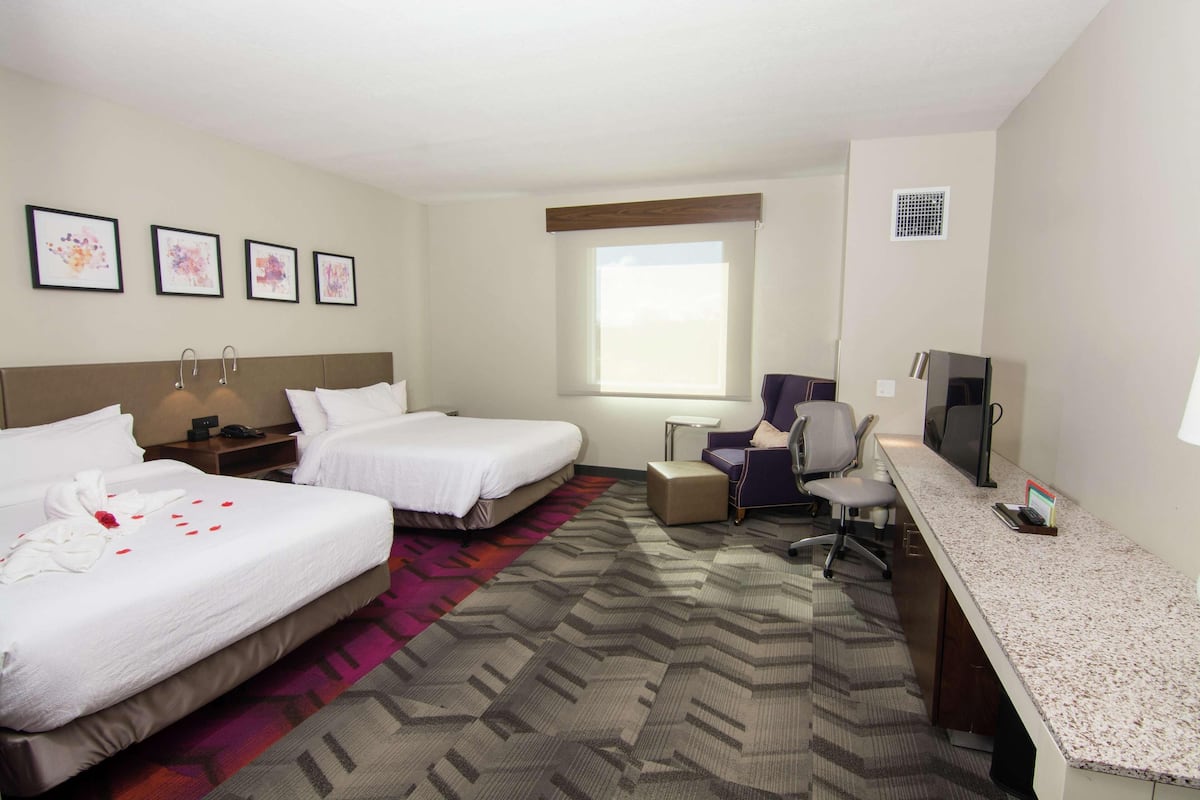 A hotel room with two beds, a desk, a chair, a TV, and a window.