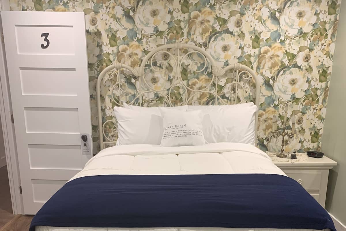 A bedroom with a bed, a floral wallpaper, a white door with the number 3, and a nightstand with a candelabra.