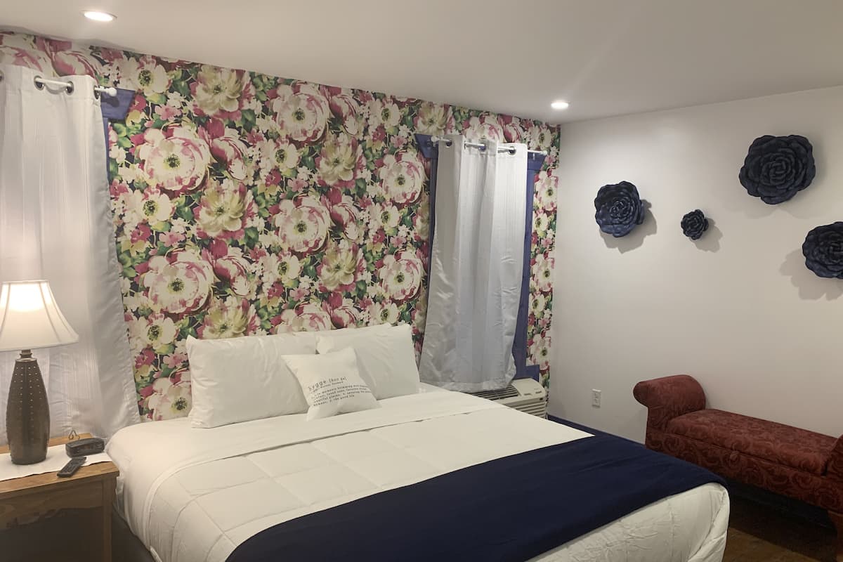 A bedroom with a floral wallpaper, a bed with white and navy bedding, a bedside lamp, a red bench, and decorative wall art.