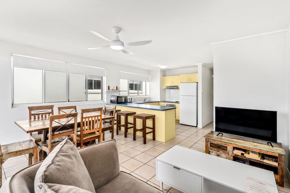 A modern kitchen with a central island, a dining area with wooden chairs, a ceiling fan, and a television on a wooden stand.