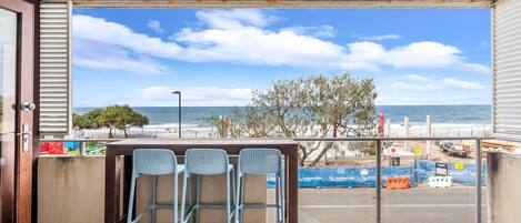 A balcony with a view of the beach, a table with chairs, and a railing.