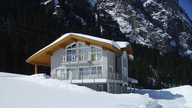 A two-story chalet with a balcony, surrounded by snow-covered mountains.