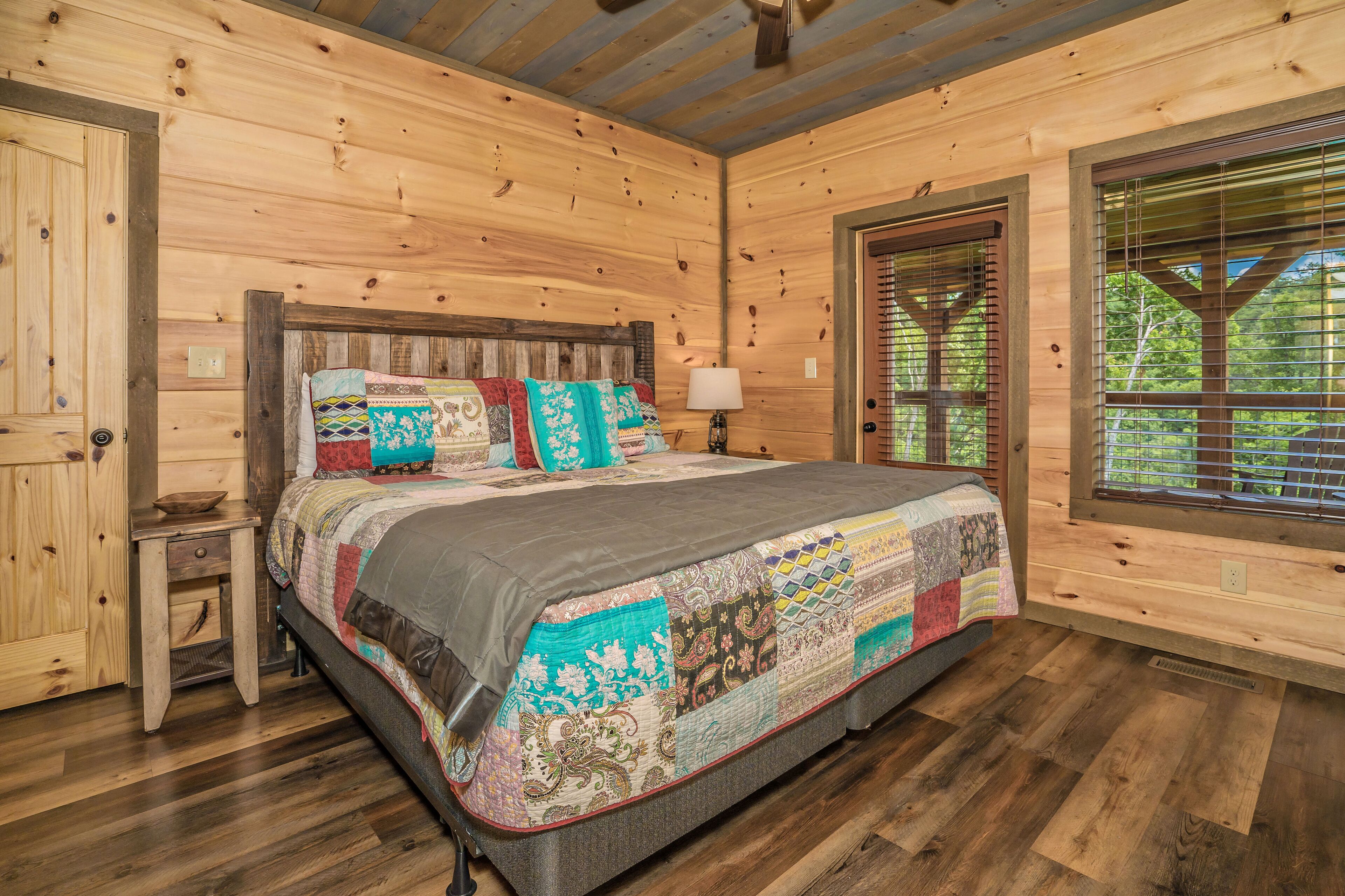 Un dormitorio de cabaña de madera con una cama grande, una mesita de noche, una ventana con persianas y una puerta.