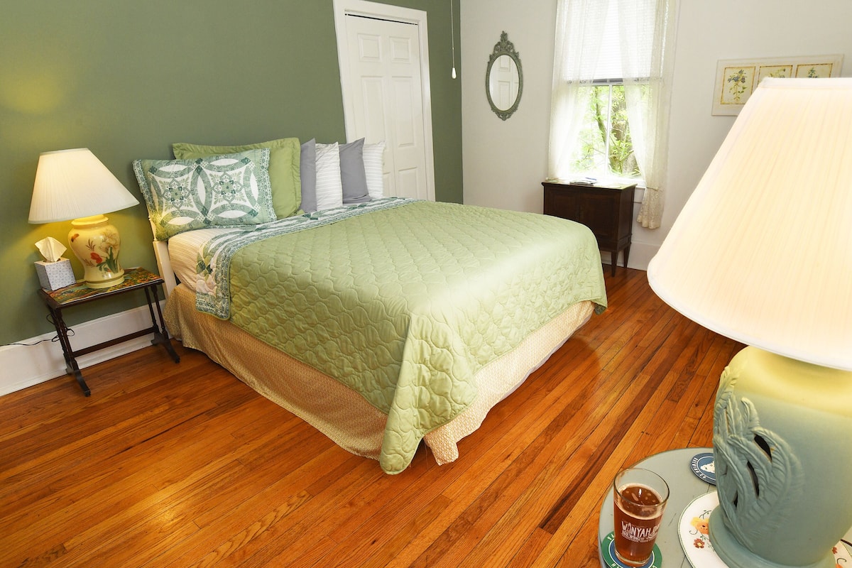 A bedroom with a bed, a nightstand, a lamp, a window with curtains, and a wooden floor.