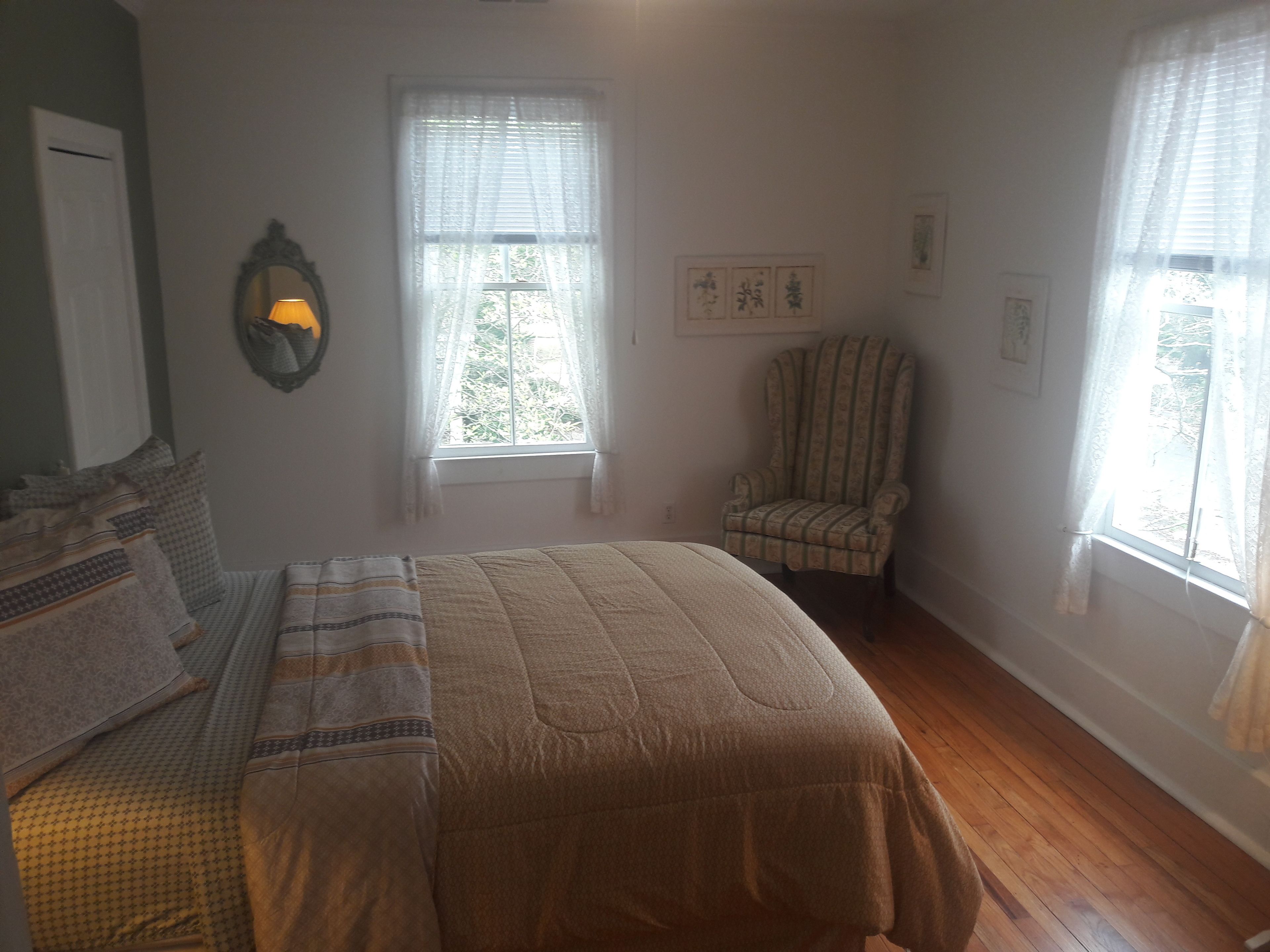 Un dormitorio con una cama, dos ventanas con cortinas translúcidas, una silla y una lámpara de pared.
