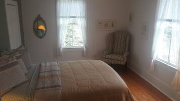 Un dormitorio con una cama, dos ventanas con cortinas translúcidas, una silla y una lámpara de pared.