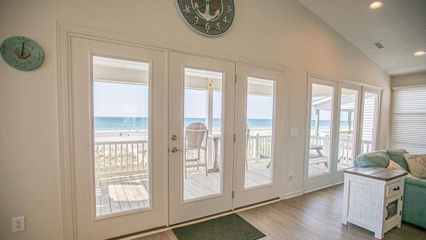 A room with a view of the beach, a clock on the wall, and a small table with a cushion. - Experience oceanfront luxury at Ohana Tides, a 5-bedroom retreat in Holden Beach. (Holden Beach)