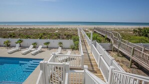 A pool area with a wooden deck leading to the beach, surrounded by lounge chairs and a white fence. - Experience oceanfront luxury at Ohana Tides, a 5-bedroom retreat in Holden Beach. (Holden Beach)