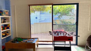 A room with a foosball table, a play area with toys, and a window with blinds.